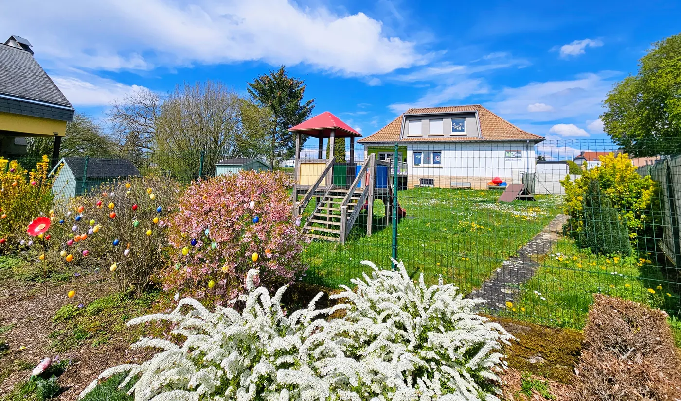 Le jardin de la crèche au printemps : massifs fleuris, cabane avec toboggan et la maison au fond